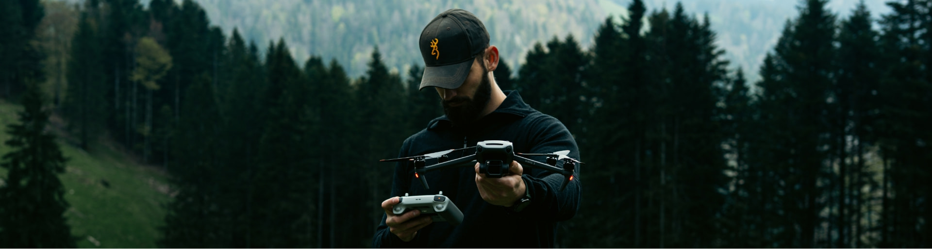 Pilote tenant un drone et sa radiocommande devant une forêt de montagne, tête baissée vers son matériel